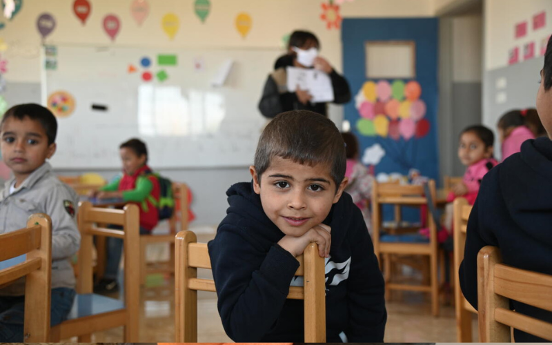 A young boy in a classroom hangs over a chair, smiling.