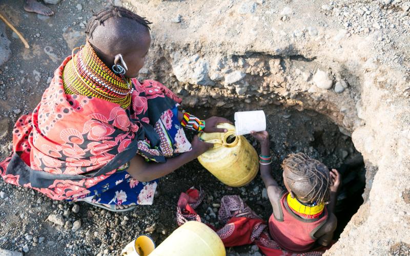 A Massai woman and child collect water from a rocky, recessed water source, pouring it into a yellow jerry can.