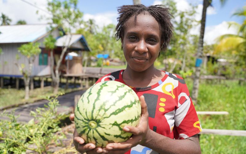 A woman wearing a colourful read shirt smiles as she holds up a melon.