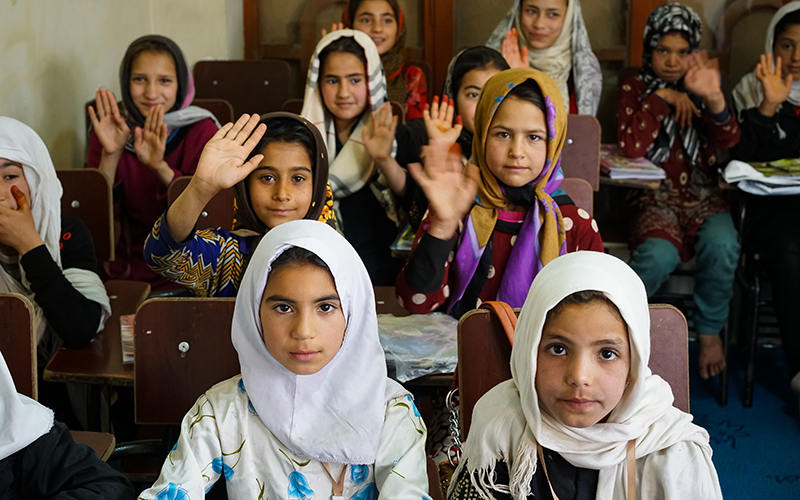A classroom of young girls in Afghanistan sit at desks and raise their hands.