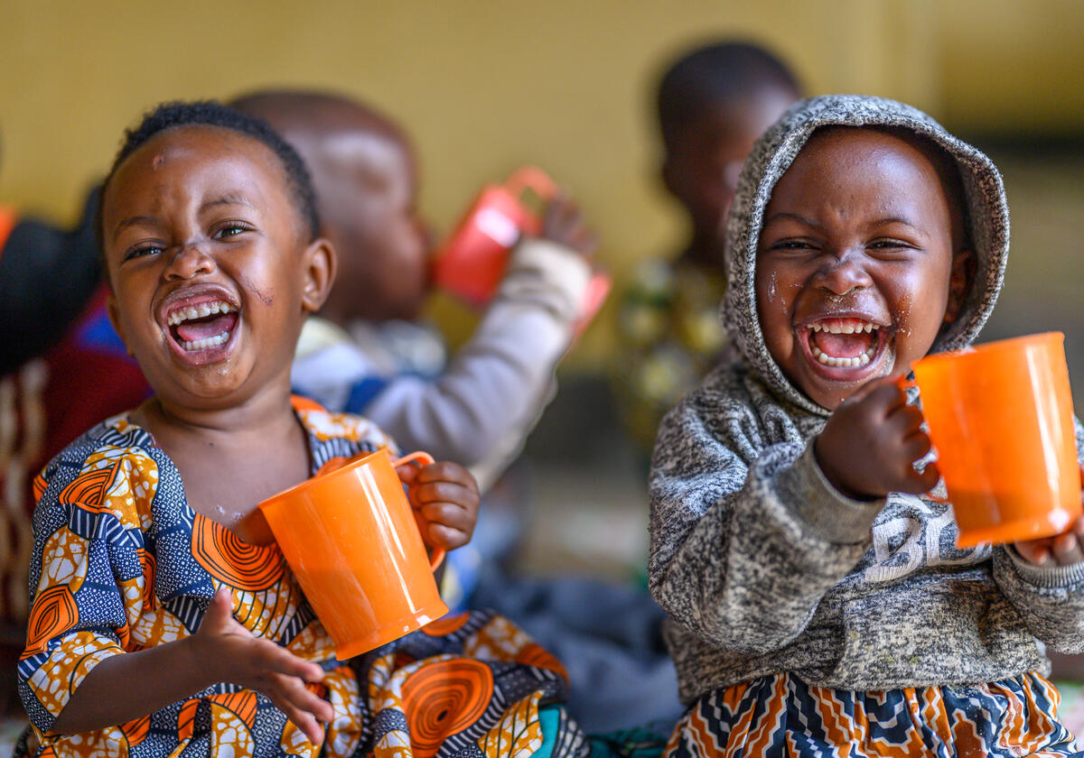 Two young children in Rwanda laugh joyously as they drink from large orange mugs.