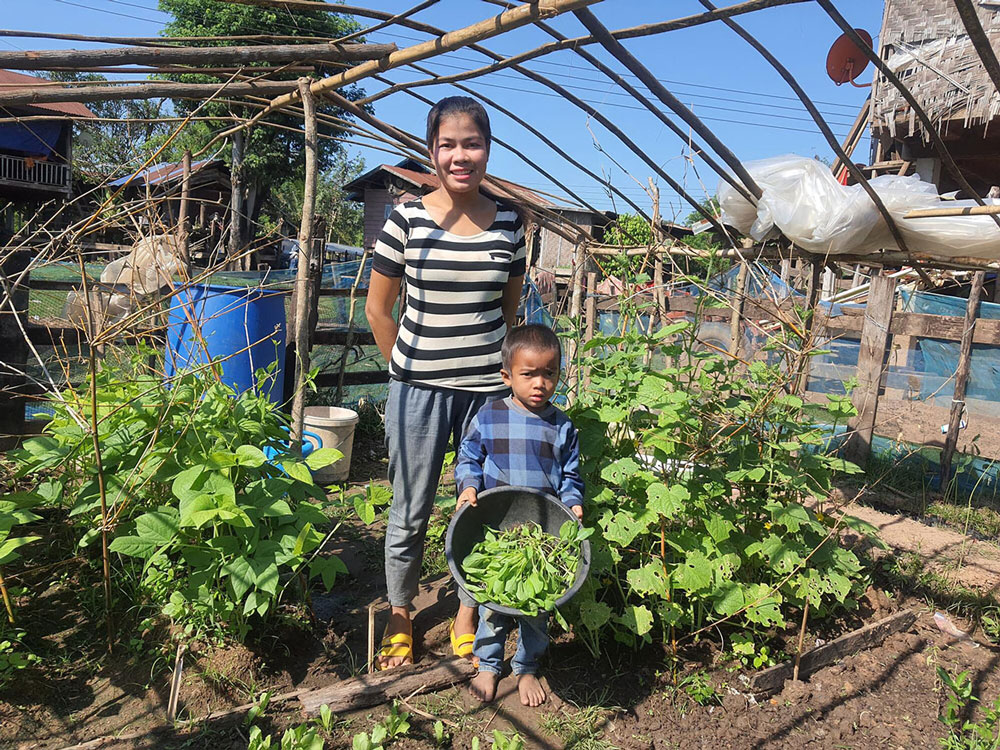 A woman stands in the middle of her garden. A makeshift roof from branches and wire protect the garden. Her young son with her and shows a bowl of fresh produce. 