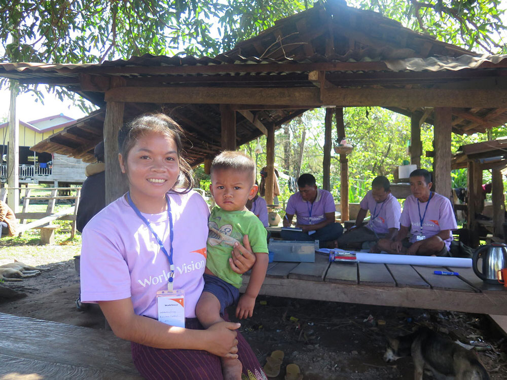 In Laos, a young woman wearing a World Vision T-shirt holds a little boy. Behind her people wearing the same shirt gather under a pavilion.