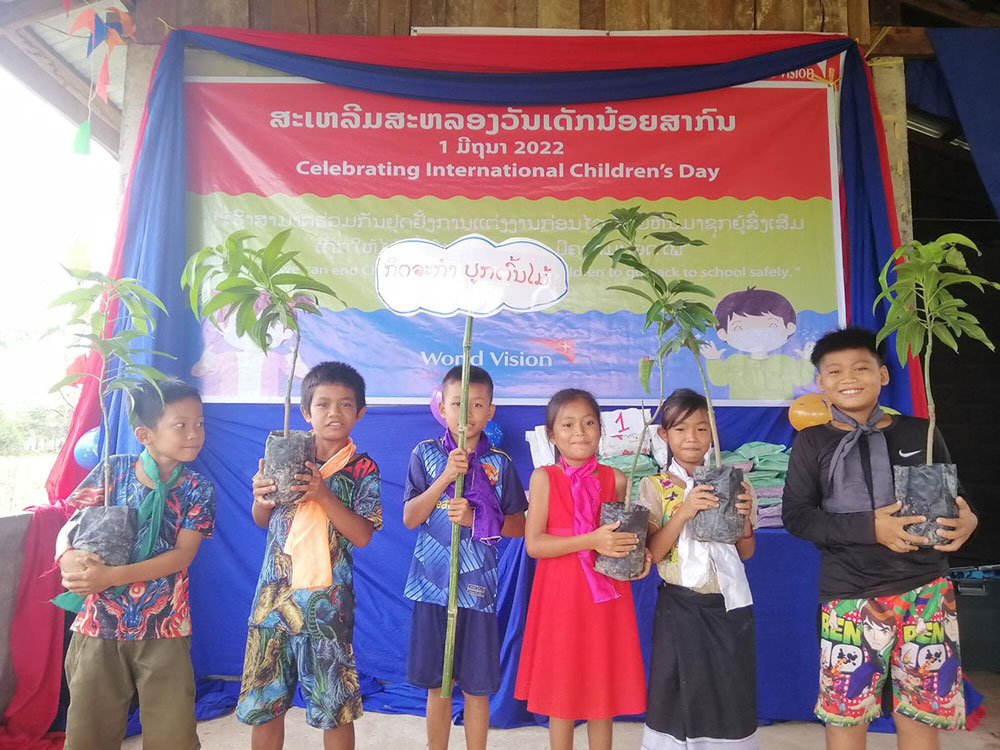 six children stand in front of a blue banner. They are holding potted mango trees. 