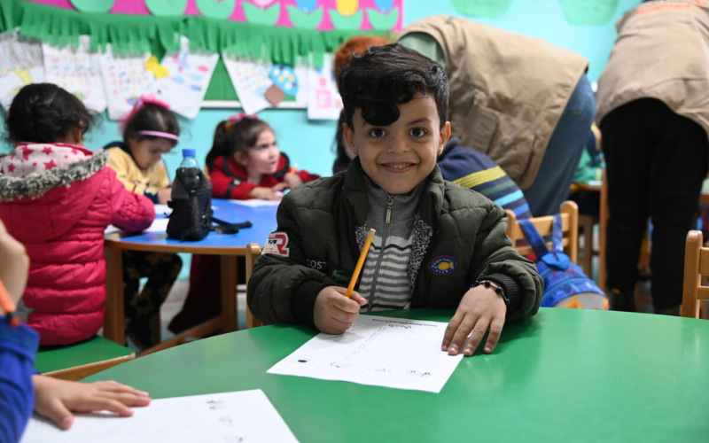 A young Lebanese boy grins at the camera, sitting in his classroom and holding a pencil.