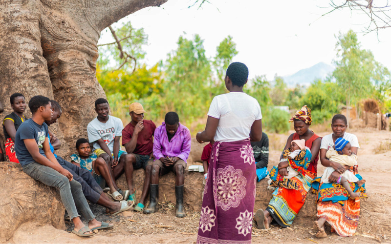 A group of young people sit under a big tree, listening to a young woman standing and speaking to them.