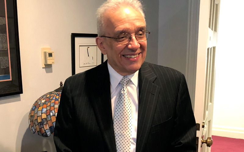 A man stands in a room, wearing a black suit and polka dot tie. He is smiling.