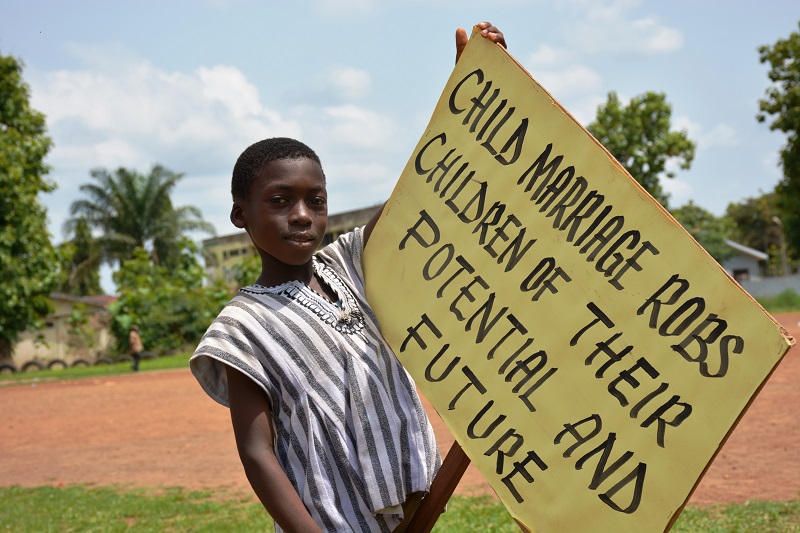 A child stands holding a sign that reads, ‘Child marriage robs children of their potential and future’.