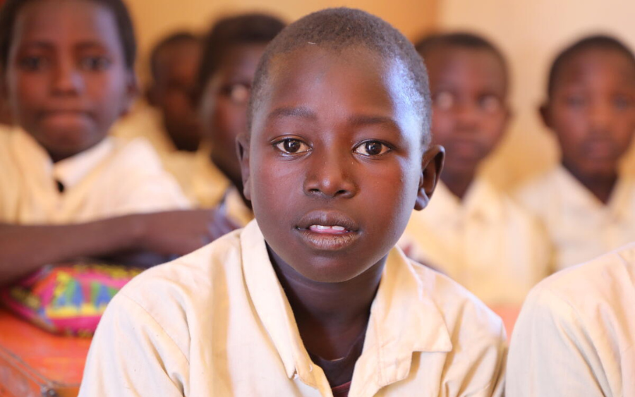 A young boy in Sudan wears a white shirt and sits in a classroom while looking directly at the camera.