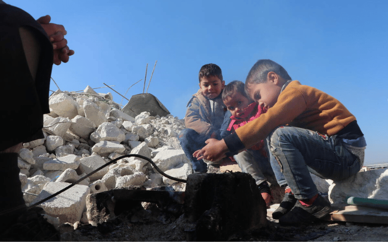 Three Syrian children squat in the rubble, warming their hands over a heater.