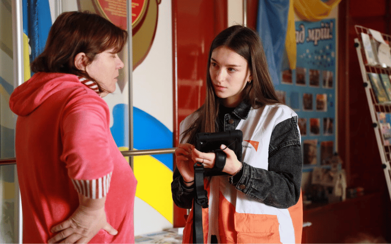 Two women speak together, with one wearing a World Vision vest and holding a computer tablet.
