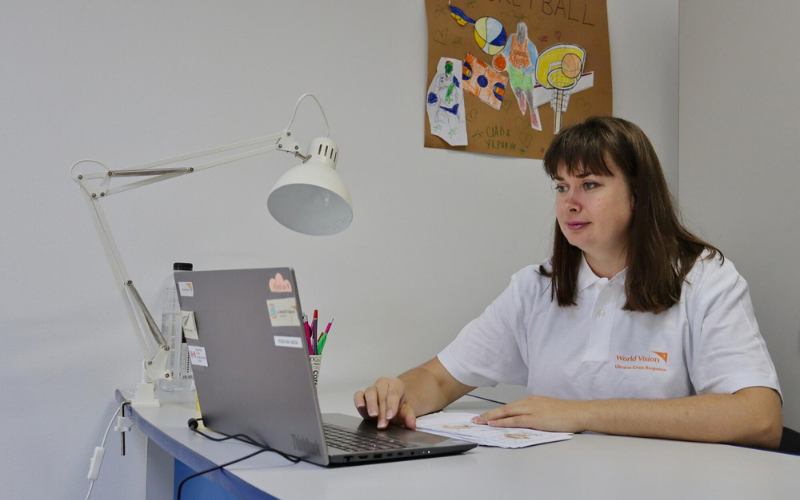A woman wearing a white World Vision polo shirt sits at a desk using a laptop.