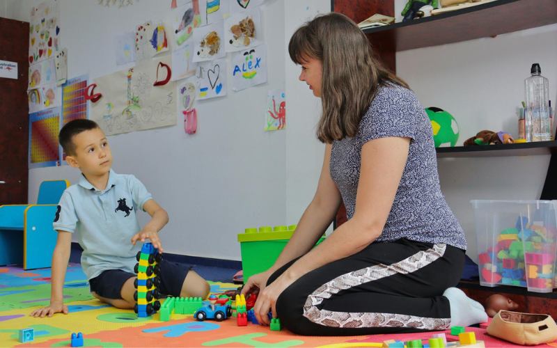 A woman and a young boy sit on a colourful floor mat and play with toys.