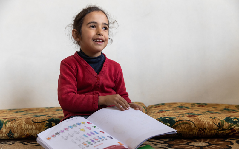 A young Lebanese girl holds a book and smiles broadly.