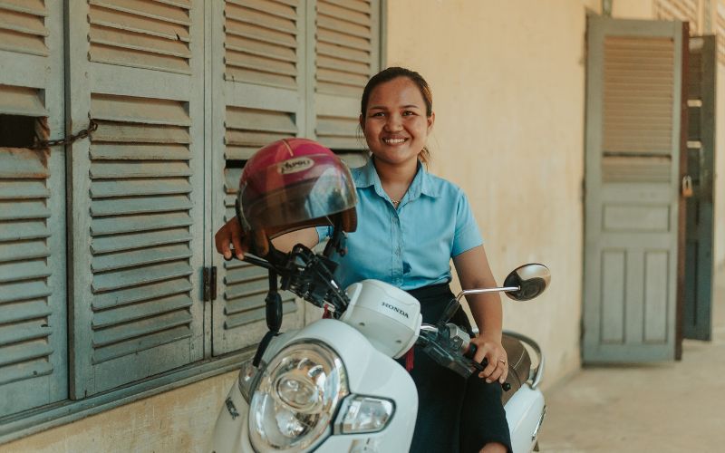 A young woman in a blue blouse sits on a motorbike outside a school building in Saang, Cambodia.