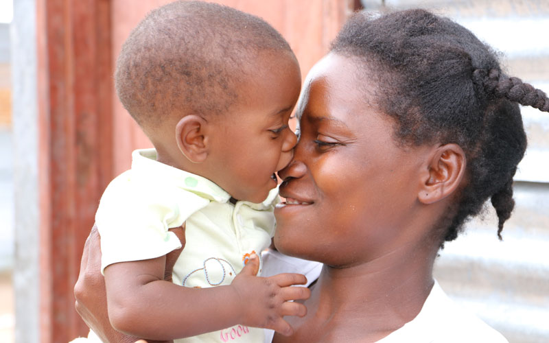 An Angolese woman holds a baby. They are smiling, and the baby appears to be going for her nose with his mouth.