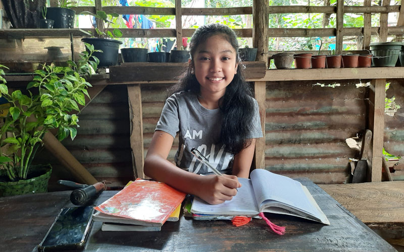 a young Filipina girl sits at a table with books open in front of her.