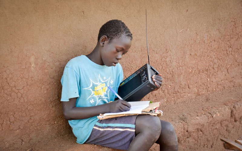 a young South Sudanese boy listens to a radio and writes in a notebook.