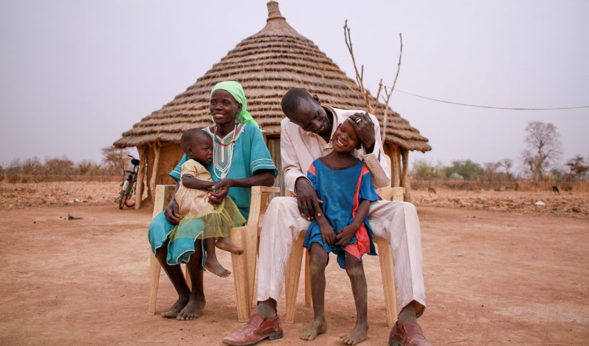 A smiling family sits in front of their hut
