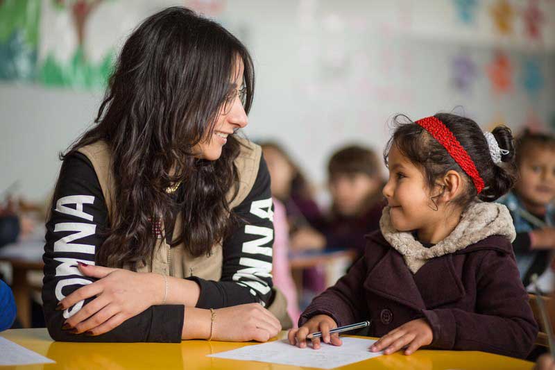 A young woman smiles at a little girl from Syria, who has pencil and paper in front of her. They are sitting together at a table in a classroom setting.