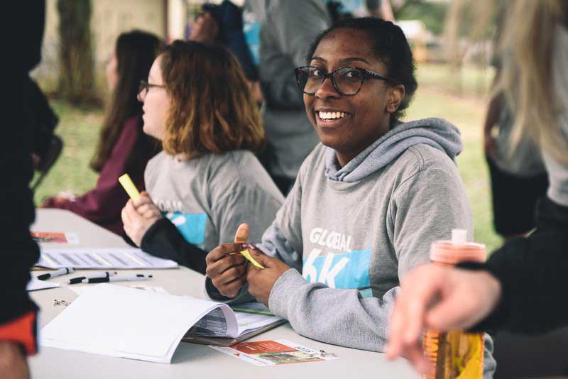A woman in a grey hoodie smiles toward the camera. She’s at a table with other volunteers. They all have papers in front of them.