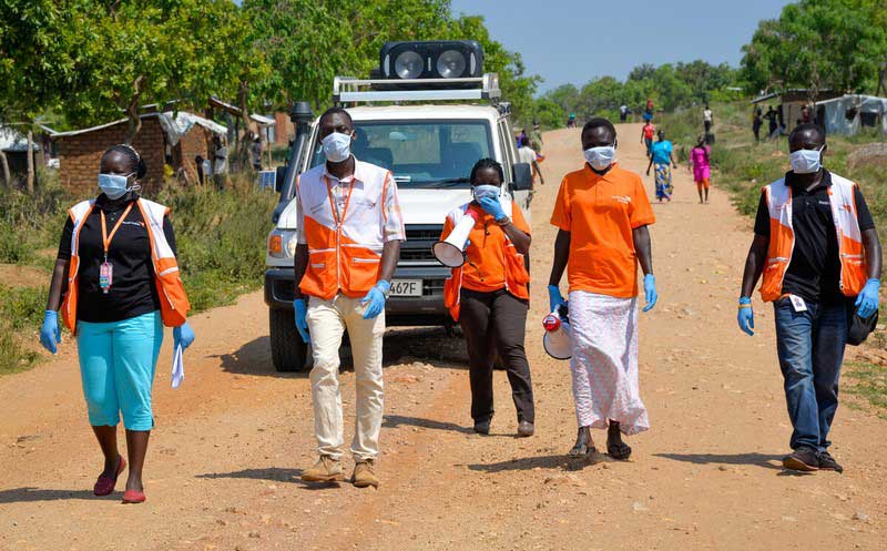 A small group of people wearing medical masks and standing six feet apart walks toward the camera. One of them has a megaphone. In the distance are huts and families.