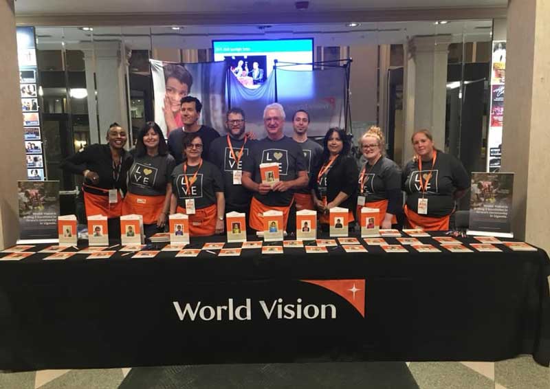 A group of people stand behind a table at a World Vision event. They are all smiling and wearing T-shirts reading “Love”. On the table are folders with photos.