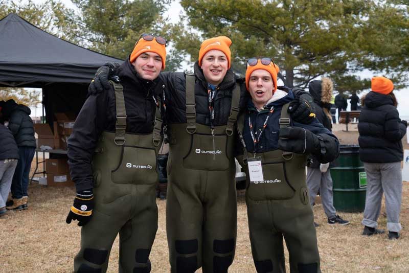Three men in orange hats, black coats and waterproof-looking pants have their arms around one another. They smile toward the camera.