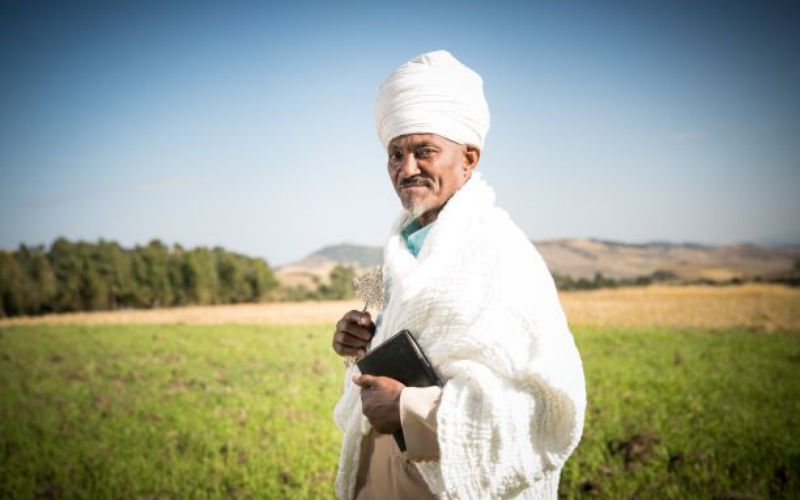 An Ethiopian Orthodox priest stands in a field wearing white linen clothing.