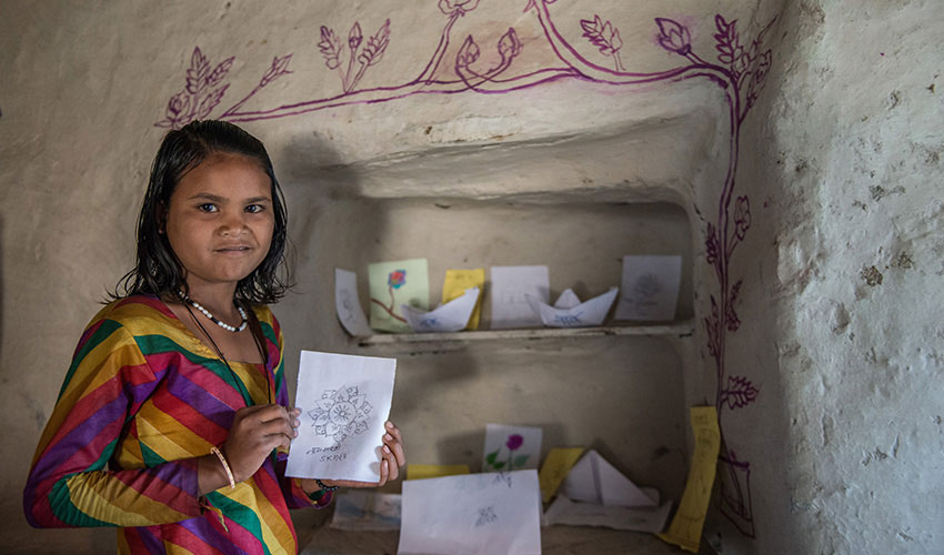 a young Indian girl stands next to her home reading corner with shelves filled with books and crafts. She smiles.