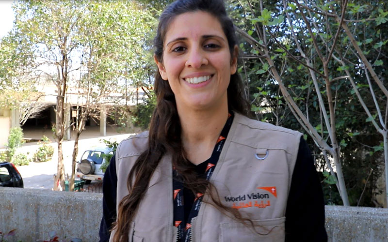 a woman smiles at the camera. She wears a beige vest bearing the World Vision logo on it