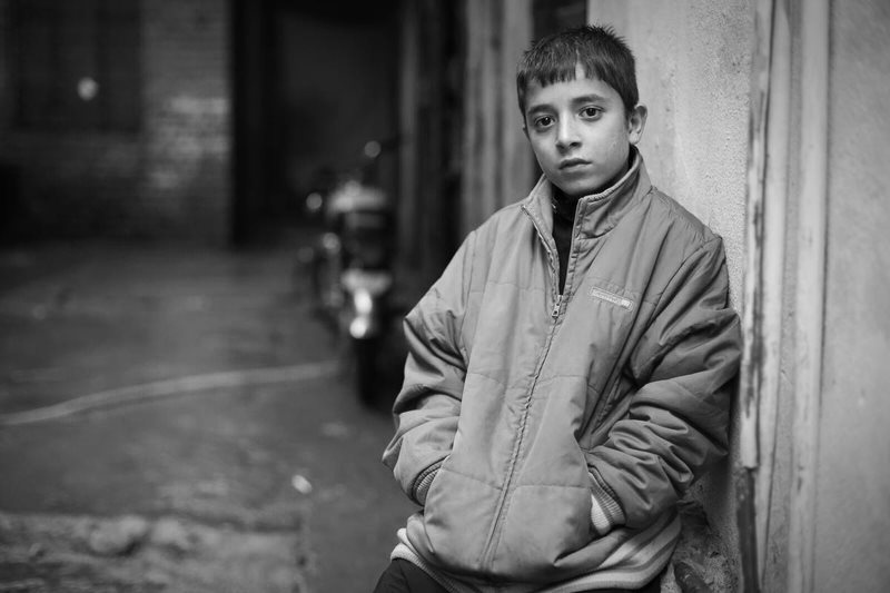 A boy from Pakistan leans against a wall in an alleyway. He looks straight into the camera.