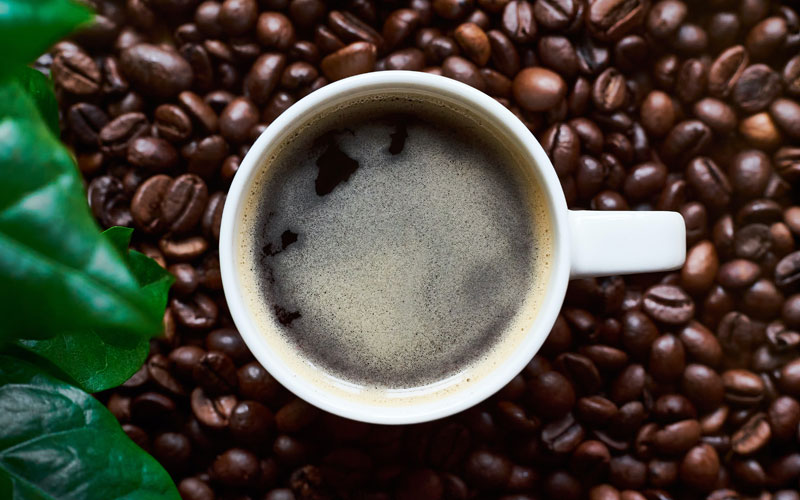 a cup of coffee on a bed of roasted coffee beans with some green leaves on the left side
