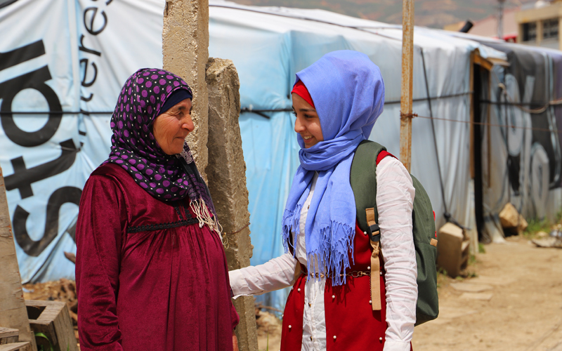 A young girl stands next to her mom. They both wear head scarves. The girl is smiling.