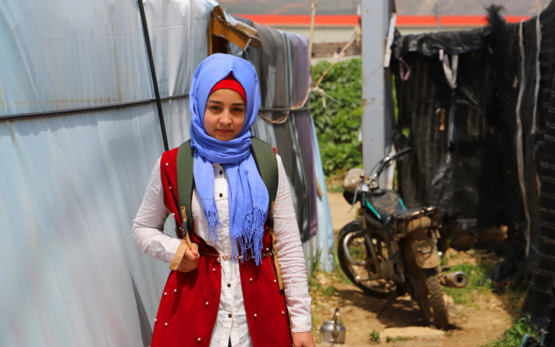 A girl stands between a tent and a motorcycle. She wears ablue head scarf, and stares straight into the camera