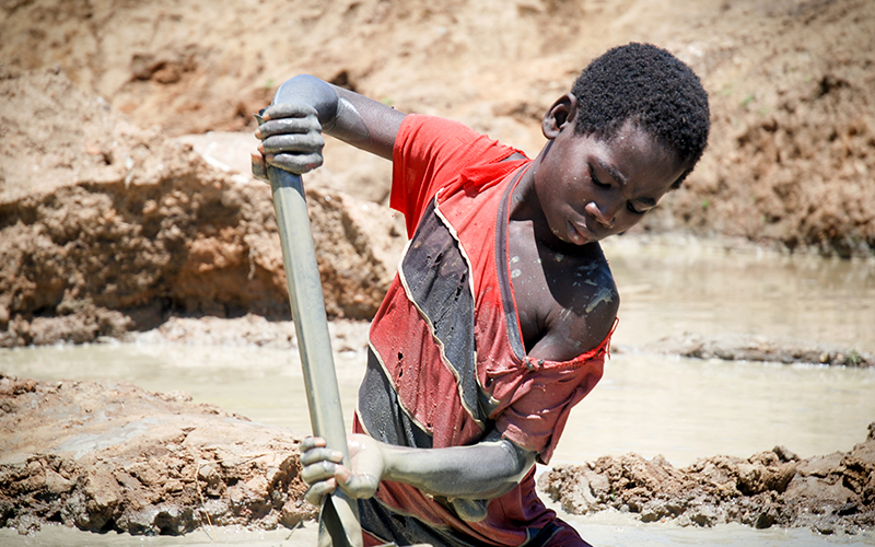 In Mozambique, an adolescent boy stands thigh-deep in muddy water, shoveling for glimmers of gold.