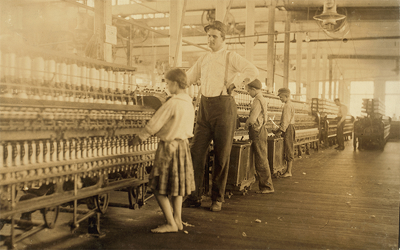 A sepia-toned image of children working on an assembly line. They are not wearing shoes. A much taller man stands over the girl in the foreground.