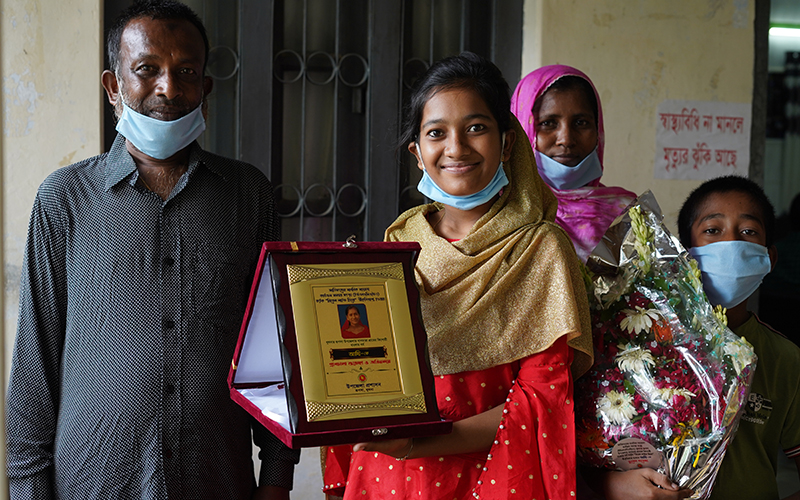 A 17-year-old girl holds up a certificate celebrating her tailoring work in the community.