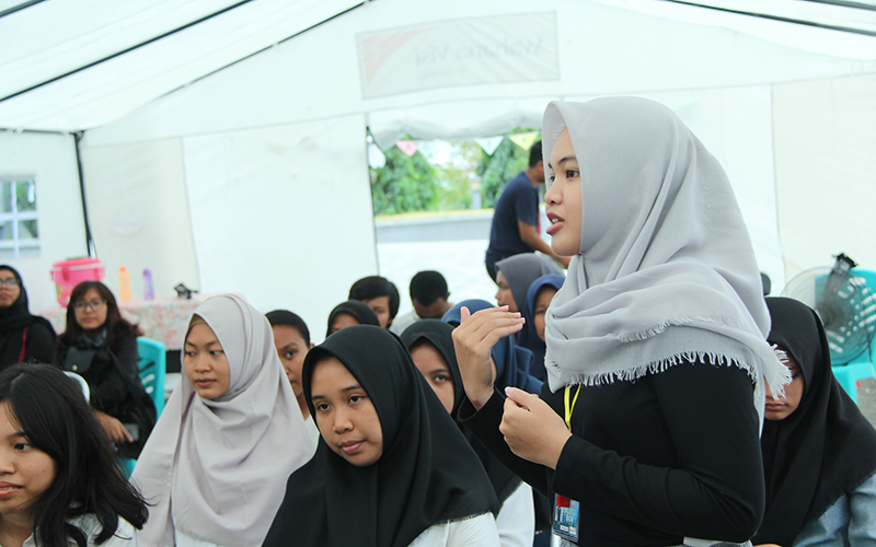 In Indonesia, teen girls wearing head scarves sit in rows listening to a girl speak.