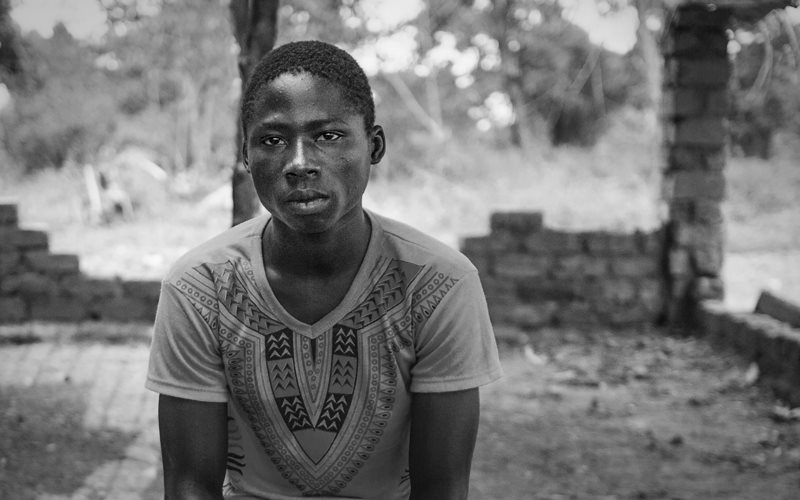 A teenager boy from the Central African Republic sits on a wall that had been demolished. He is staring at the camera, with a serious face.