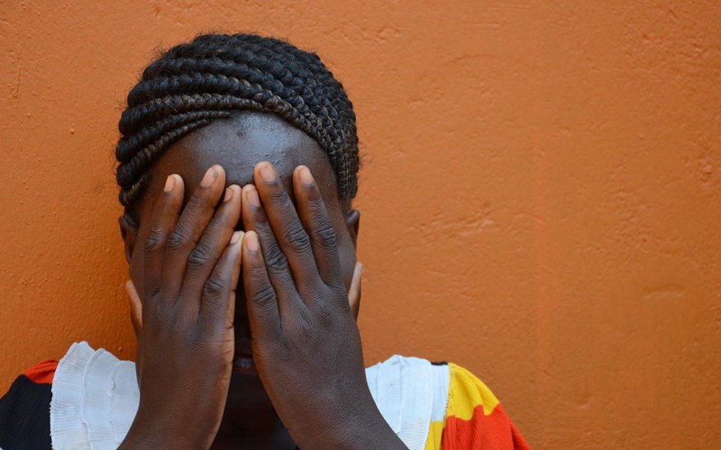 A young black girl stands against an orange wall, with her hands covering her face.
