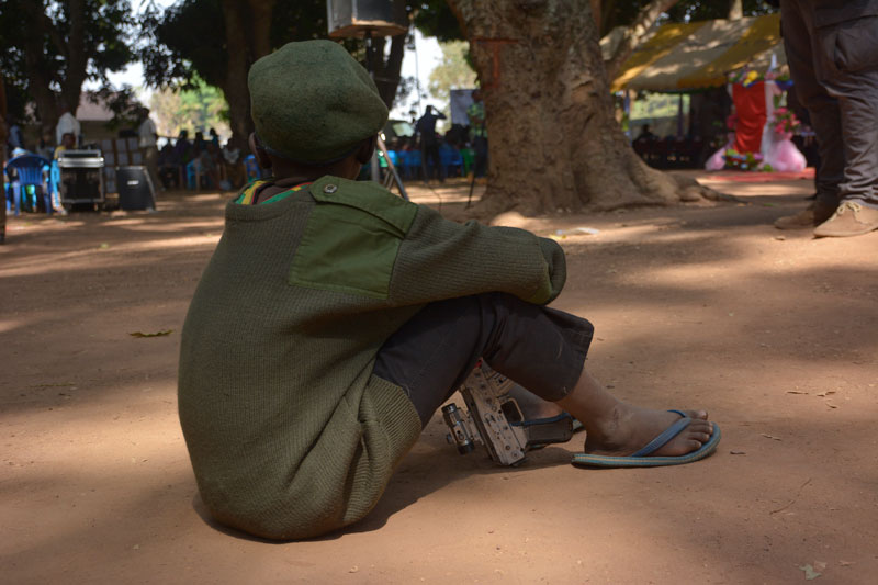 Young child soldier boy sits on a dirt ground holding a machine gun with his back to the camera.