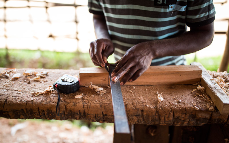 a young man from DRC measures a piece of wood with a ruler in a workshop.