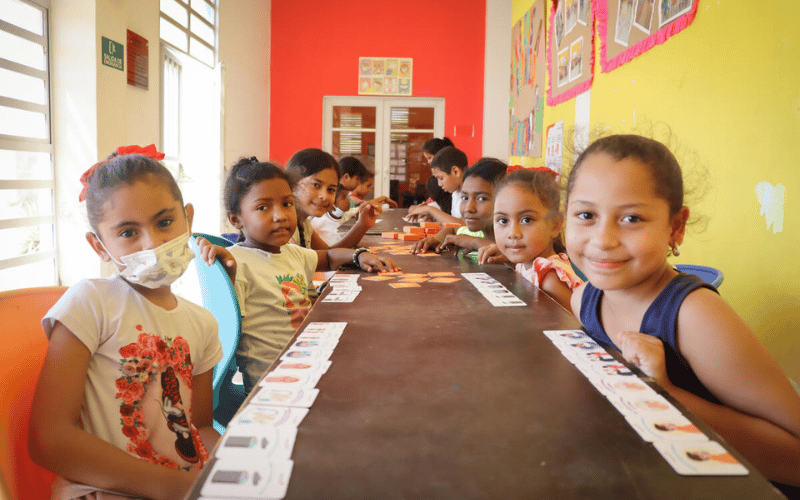 A group of young girls sit together at a long table in a classroom, with cards in front of them.