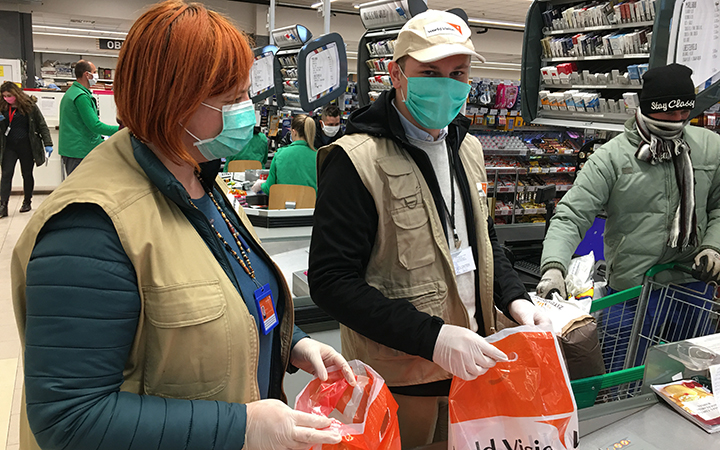 A woman and man wearing protective face masks and gloves help bag groceries at a checkout counter in a supermarket.