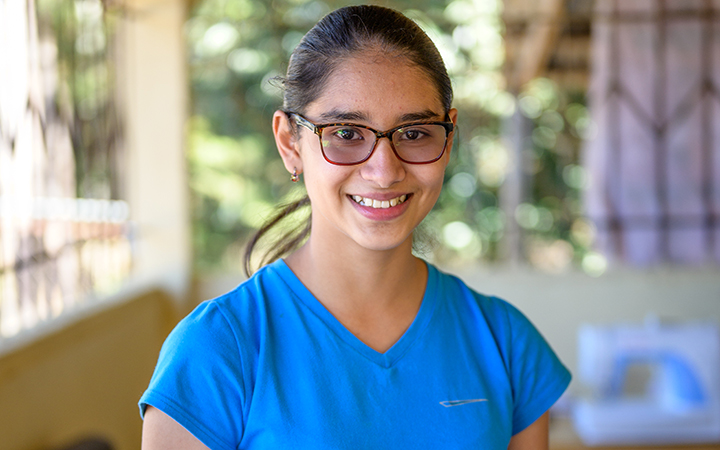 A teenaged girl in a blue top and glasses smiles into the camera.