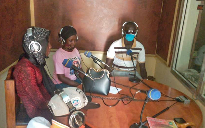 Three people sit around a table in a radio studio with mics at their mouths.