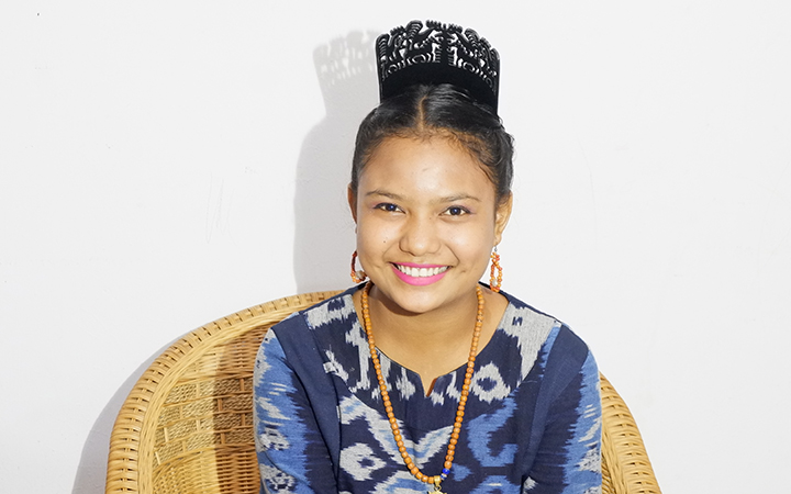 A teenaged girl in a colourful blue dress and jewelry smiles into the camera while sitting on a rattan chair.