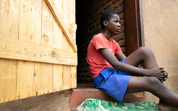 A young boy in a red T-shirt and blue shorts sits in the doorway of his home
