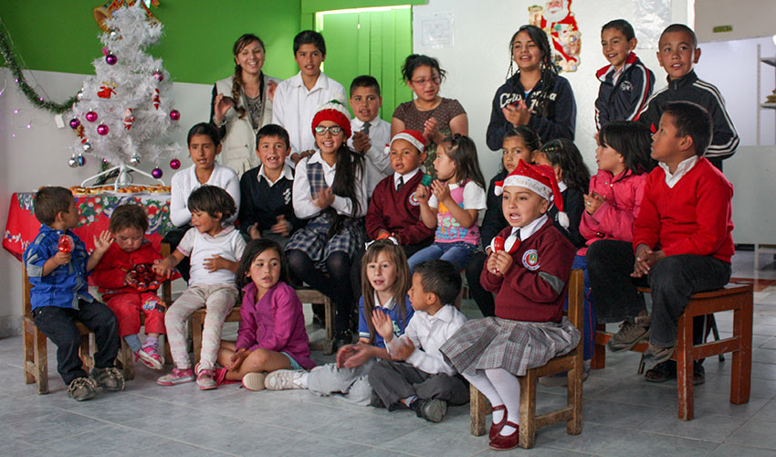 In Colombia, a group of kids of varying ages sit and stand around a white Christmas tree. They are clapping and singing.